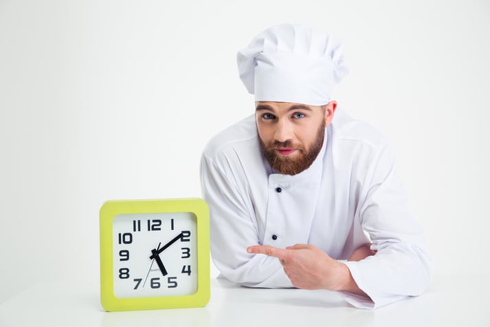 Portrait of a chef cook leaning on the table and pointing finger on clock isolated on a white background